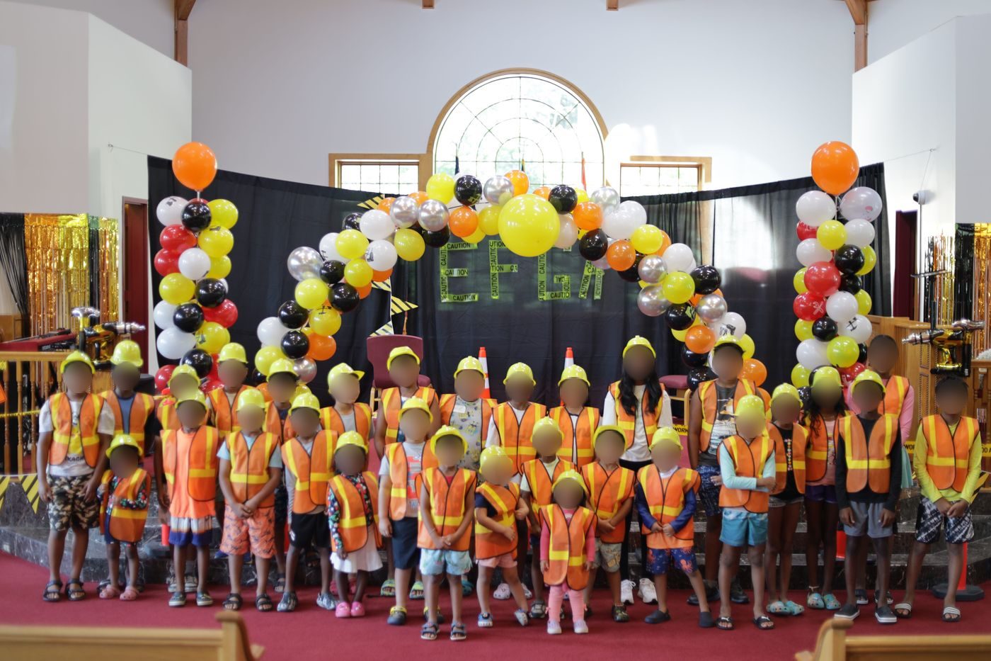 Children in orange vests and yellow hard hats gathered on the sanctuary stage under a balloon arch at Vacation Bible School